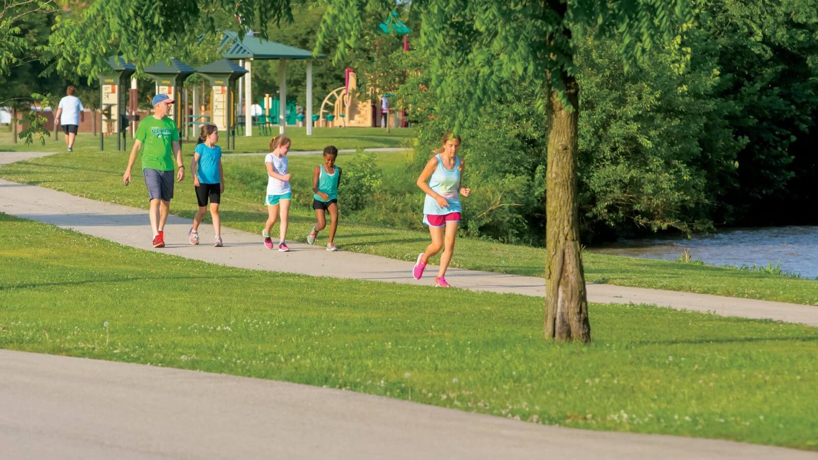 Woman jogging near park on greenway trail with father and kids behind her in Yorktown, Indiana